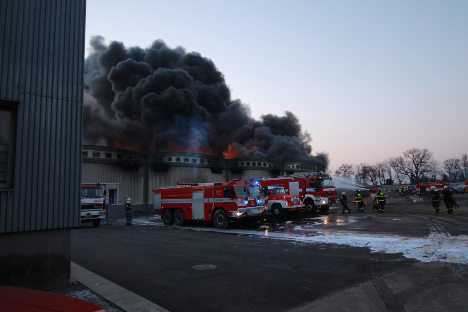 SČK_Požár mrazíren v Mochově_z budovy stoupá hustý černý kouř, před ní stojí hasičská technika a hasiči, kteří zasahují.jpg