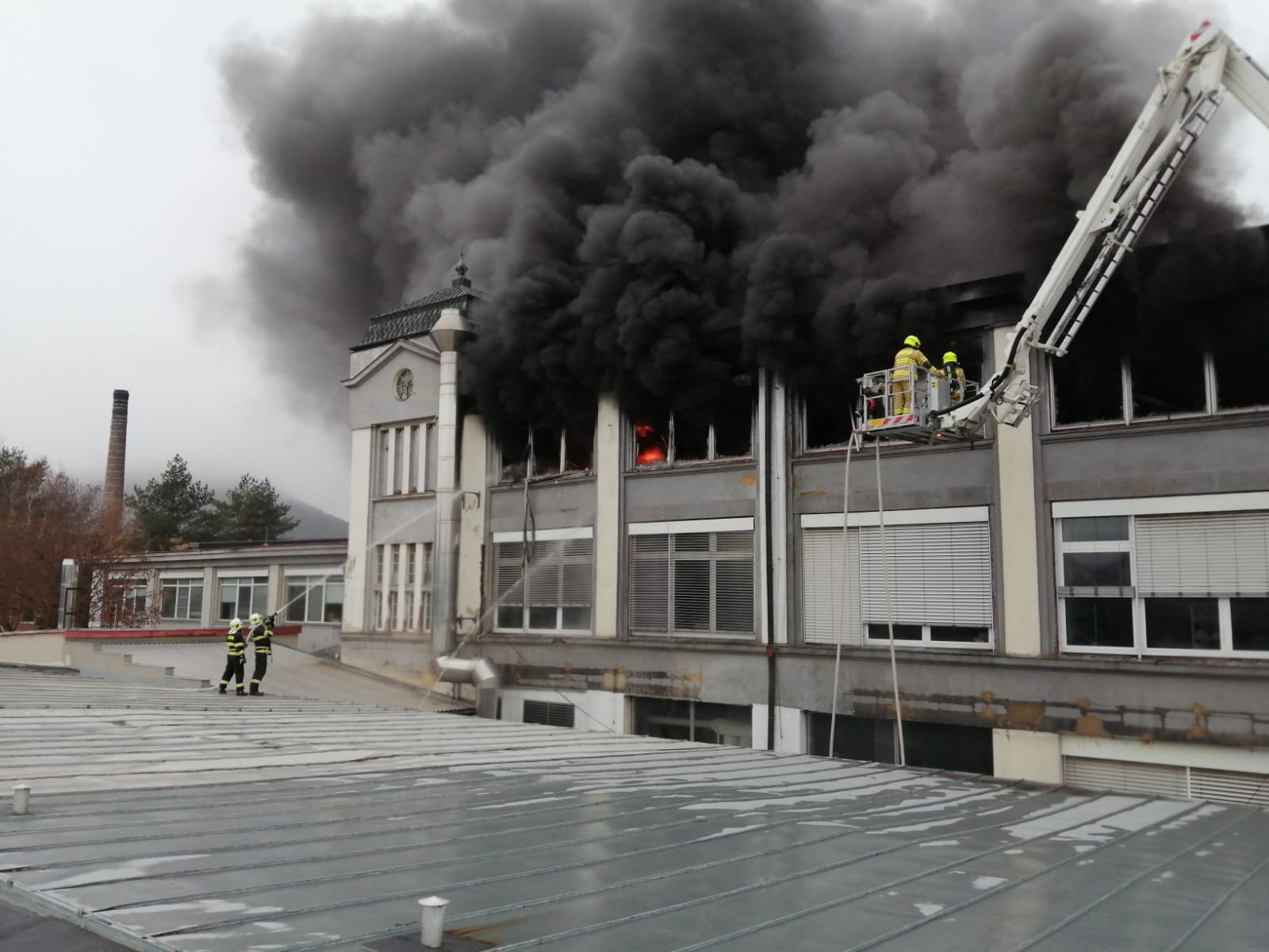 ÚLK_požár haly s plastovými modely v Mostě_hasiči z plošiny i ze střechy níže položené haly hasí hořící silně zakouřenou halu.jpg