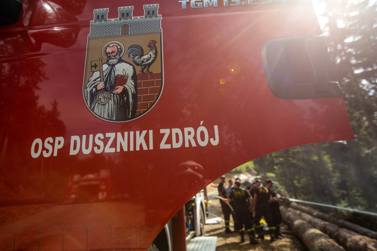 KHK_cvičení_společný zásah českých a polských hasičů při lesním požáru_pohled na auto polských hasičů a v pozadí hasiče stříkající vodu do lesa.jpg