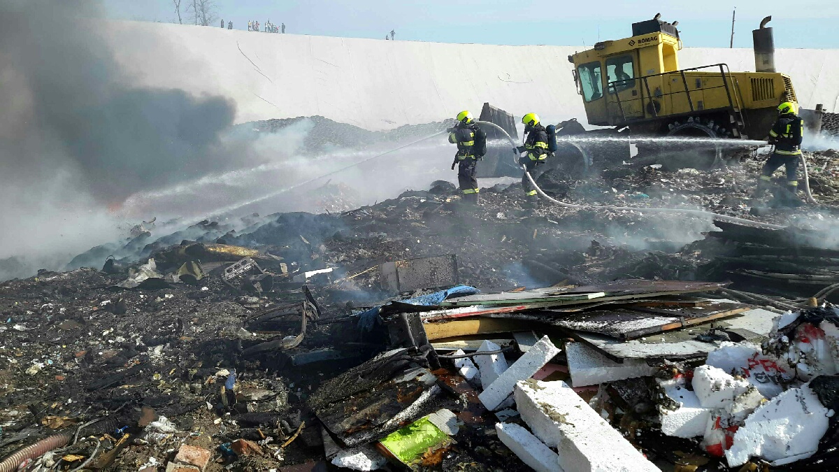 JMK_Požár skládky odpadu na Brněnsku_pohled na 3 hasiče kteří pomocí 2 proudů hasí oheň, v pozadí těžká technika rozhrabává odpad.jpg